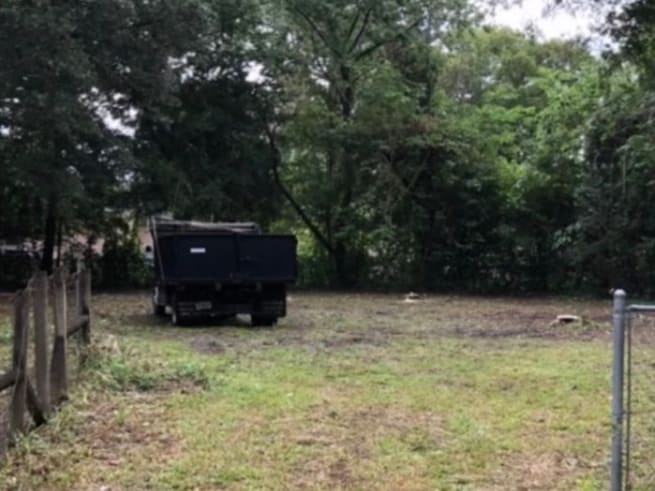 After — the same lot cleared to open grass, with a black dump trailer parked at the tree line.