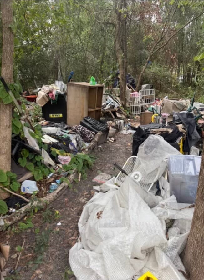 Before — a pile dumped in the woods: a cabinet, a crib, storage bins, tarps and bagged debris.